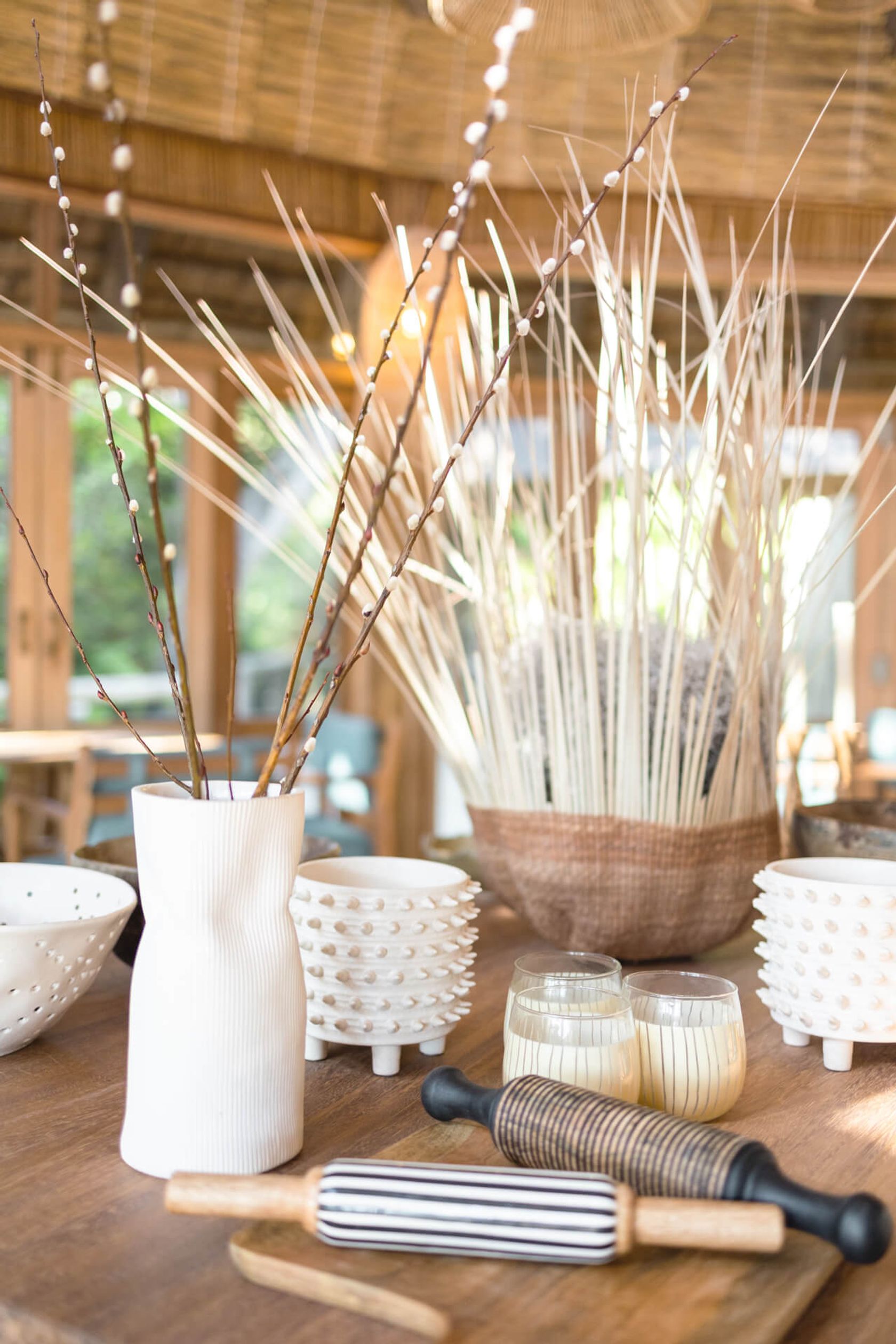 Tabletop still life with a white ribbed vase of catkin branches, studded bowls, striped glasses and a wooden rolling pin.