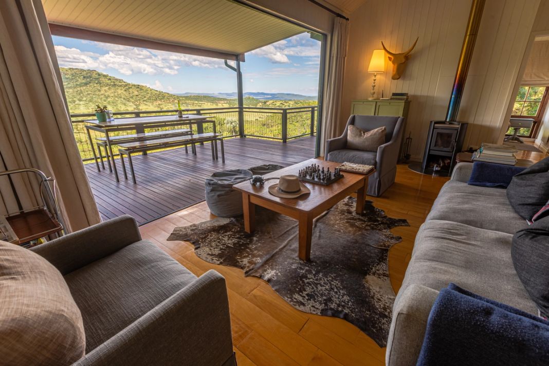 A cottage lounge opening to a deck with a dining table, looking over green hills to a distant dam under a cloudy sky.