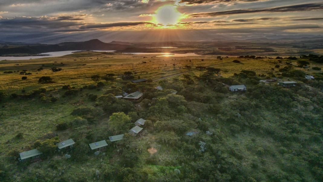 An aerial view at sunset of the lodge and its cottages set among green bush, with a dam and mountains beyond.