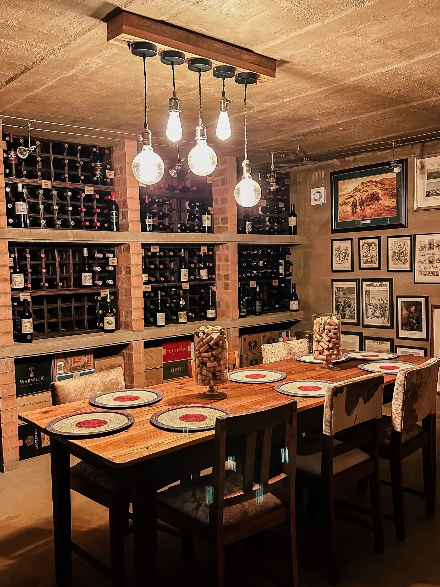 Brick wine racks full of bottles under hanging edison bulbs, beside a set table with cork jars and a wall of framed historical prints.