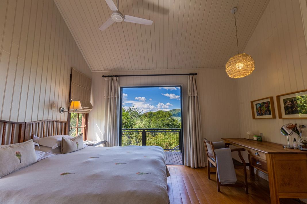 A bedroom with a vaulted white ceiling, an embroidered cream duvet, a wooden desk with mirror and sliding doors to a green view.