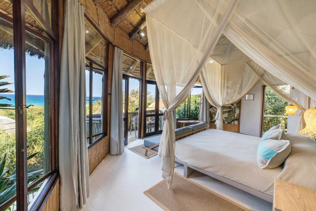 Four-poster bed with mosquito net and a blue daybed, beside corner windows framing the sea over green vegetation.