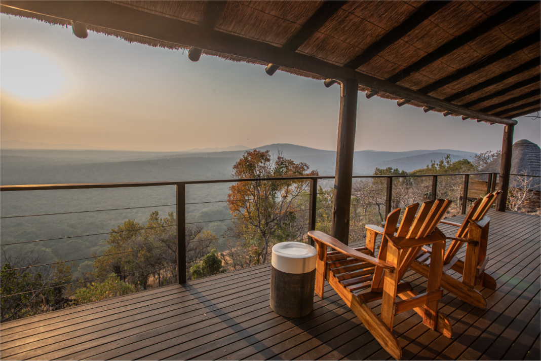 Two wooden deck chairs under a thatched roof overlooking a hazy valley at sunset.