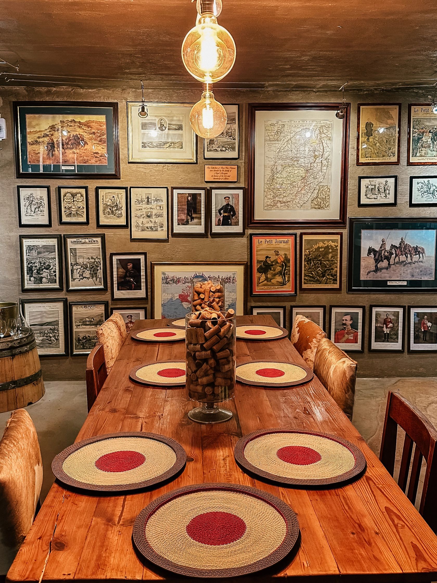 A wooden table set with woven red-centred placemats and a glass jar of wine corks, below a wall of framed anglo-boer war prints and maps.