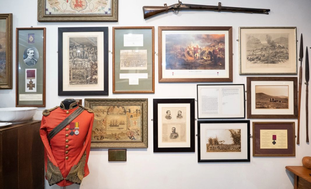 A museum wall of framed Anglo-Zulu War prints, a red military tunic, medals, and mounted weapons.