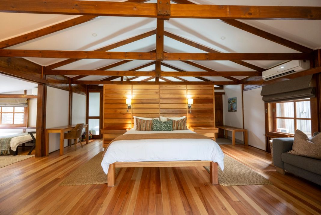 A luxury safari lodge bedroom featuring a large bed with wooden headboard, exposed ceiling beams, polished wood floors, and modern furnishings.