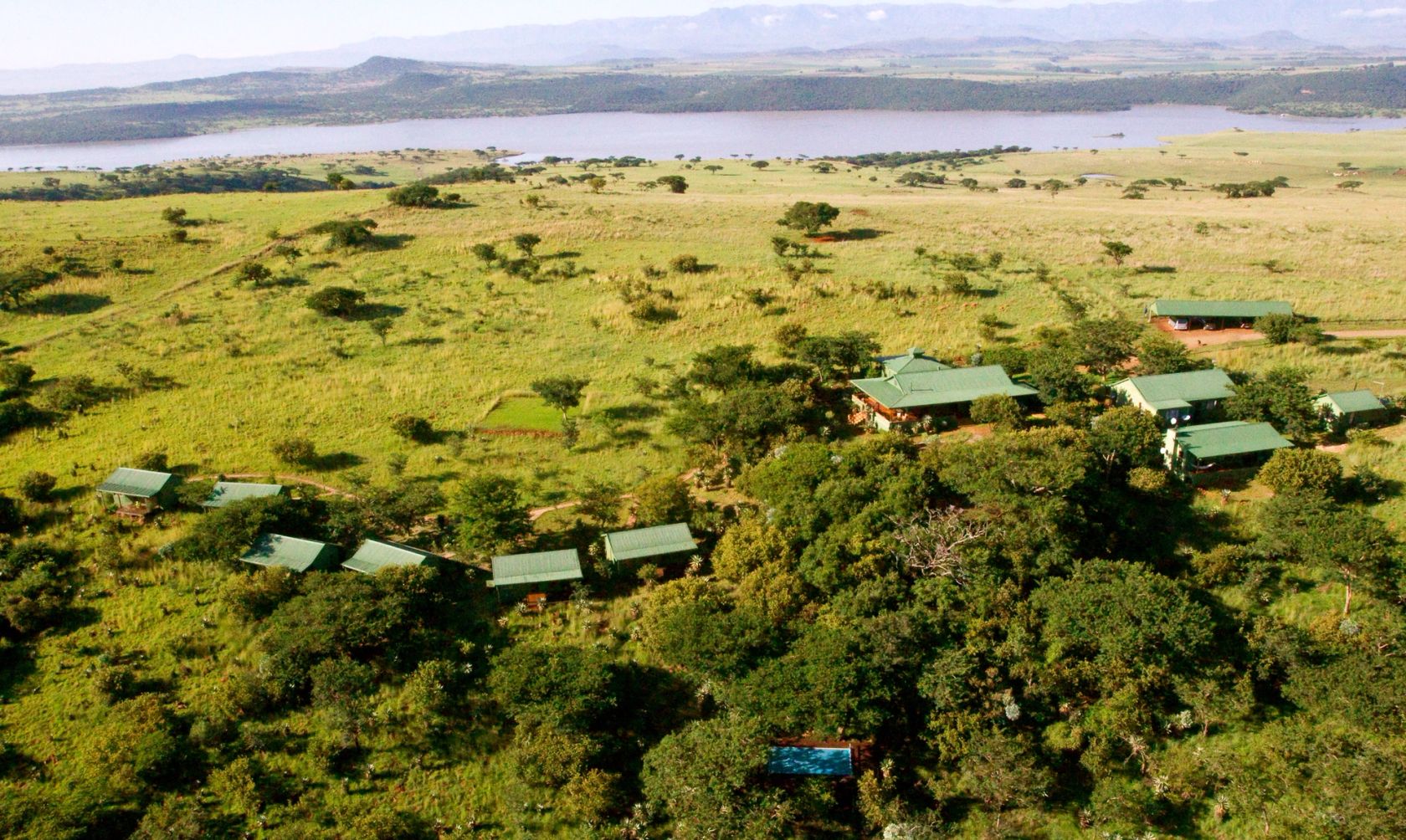 Aerial of the lodge's scattered green-roofed cottages and a small pool among trees, with Spioenkop dam and hills in the distance.