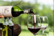 A hand tips a labelled bottle to pour red wine into a glass beside more glasses against green foliage.