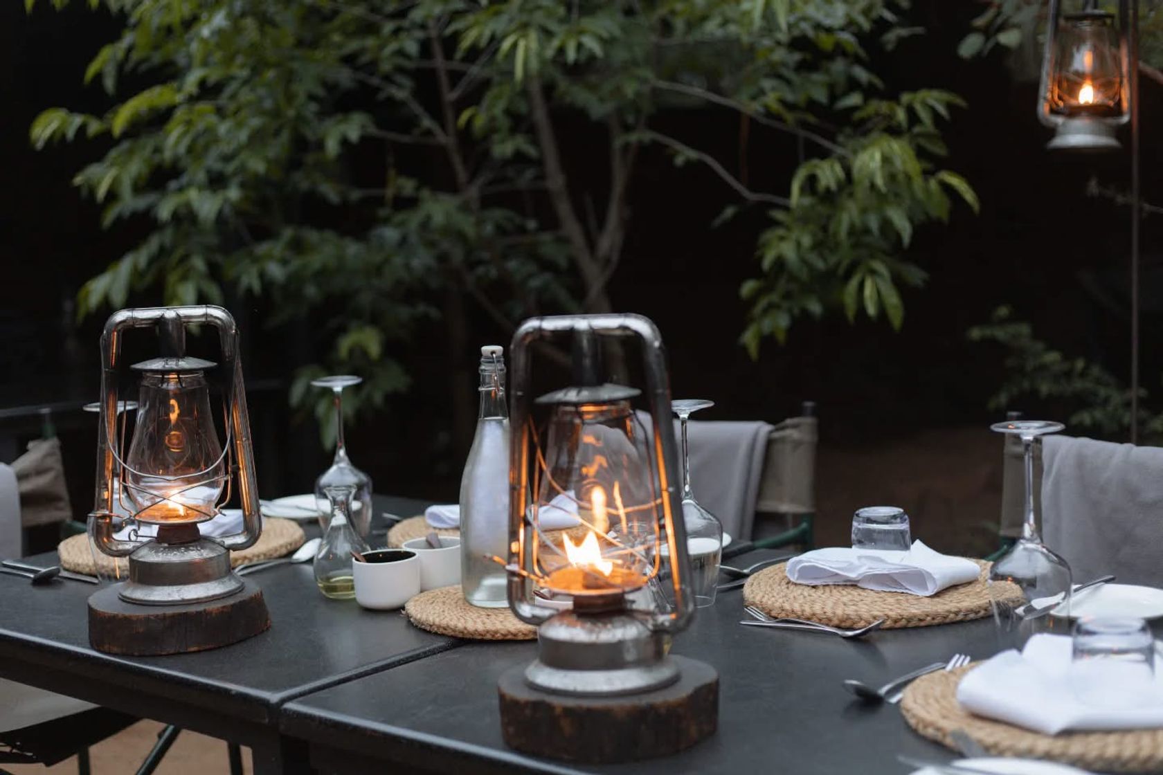 Vintage lanterns, glassware and woven placemats set on an outdoor dining table at dusk.