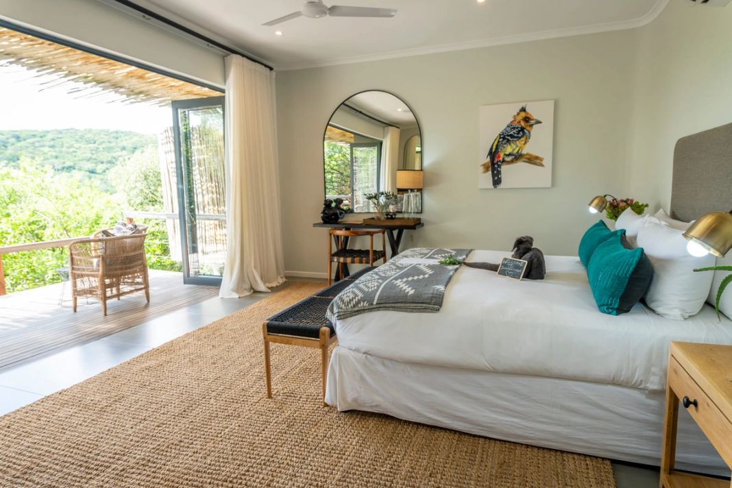 A king bed with teal cushions and a bird painting faces open glass doors leading to a deck over a green valley.