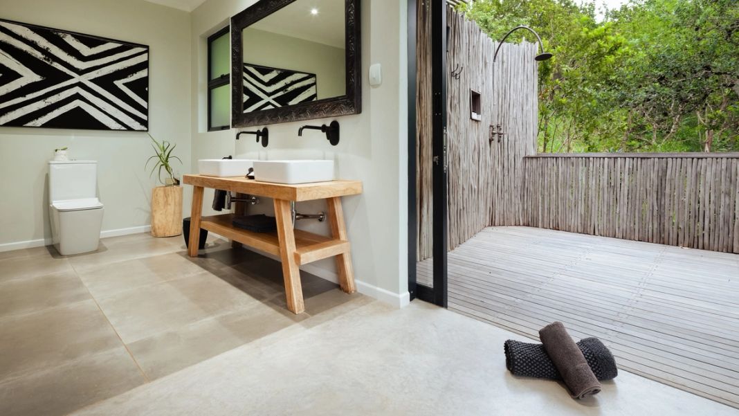 A suite bathroom with twin basins on a wood vanity, a toilet, monochrome artwork and an adjoining reed-walled outdoor shower deck.