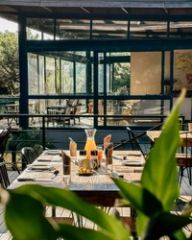 A breakfast table set on the deck with a juice carafe, framed by foliage and the glass dining room behind.