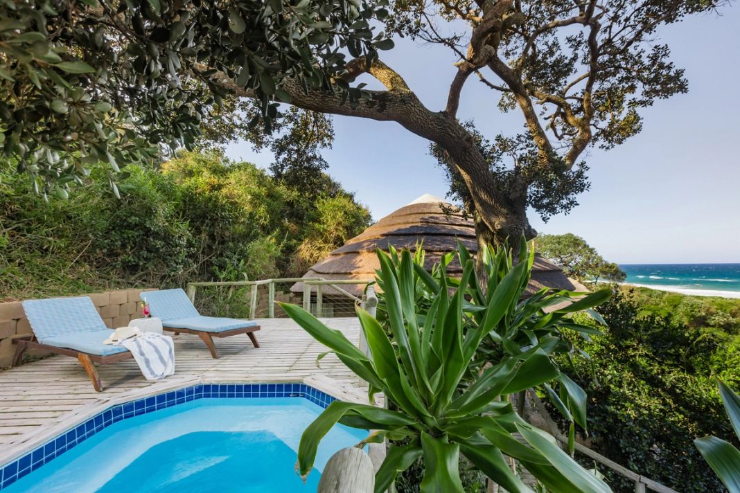 A private plunge pool and two loungers on a wooden deck, with a thatched roof and the ocean beyond.