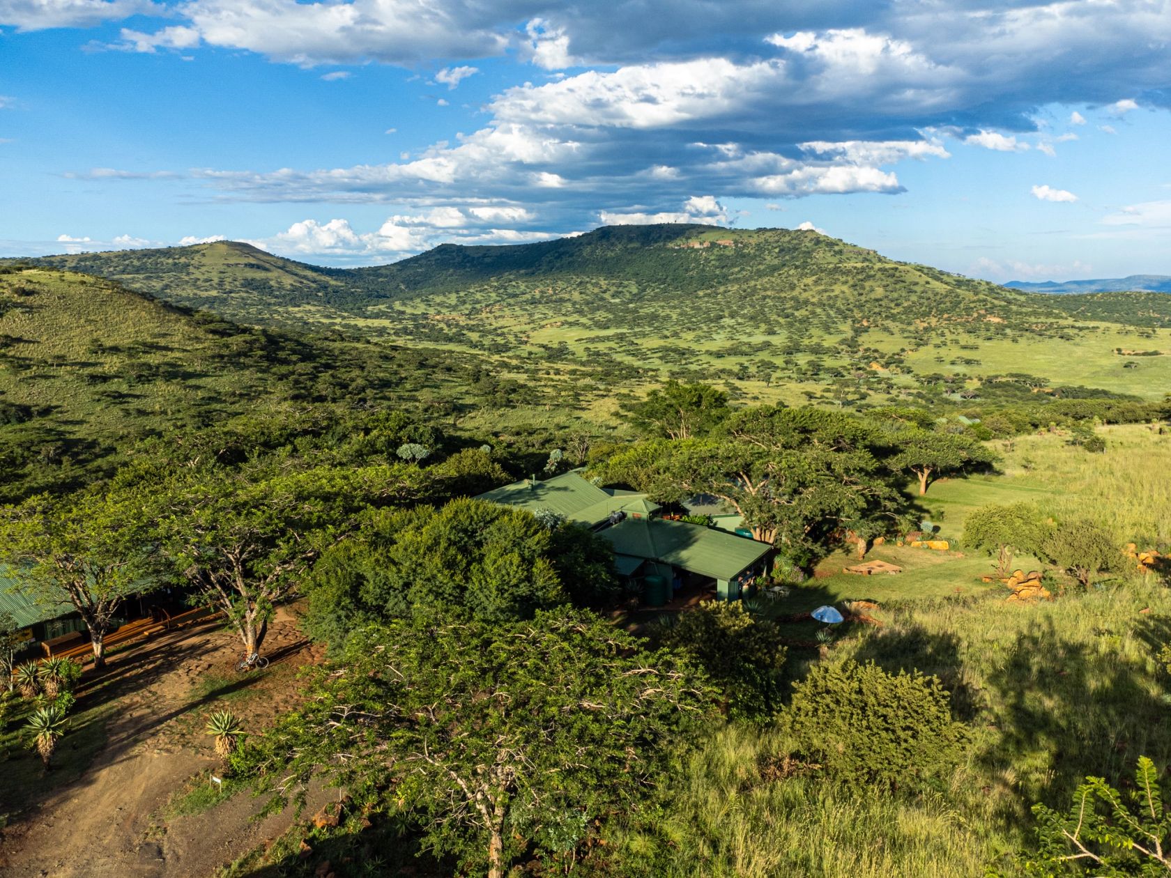Aerial view of the lodge's green-roofed buildings set among trees, looking across a green valley toward the hills below Spioenkop.
