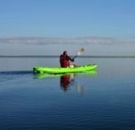 A single paddler in a red life vest sits in a green kayak on calm reflective water.