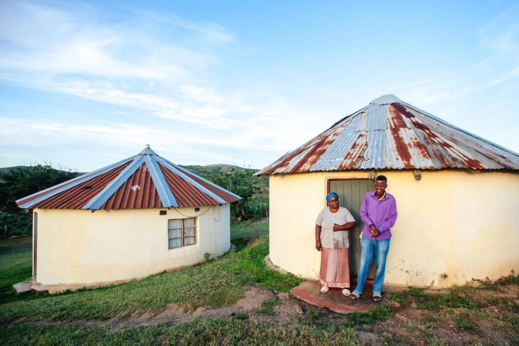 Two white-walled rondavels with corrugated roofs in the Mpembeni community, a man and a woman by the door.