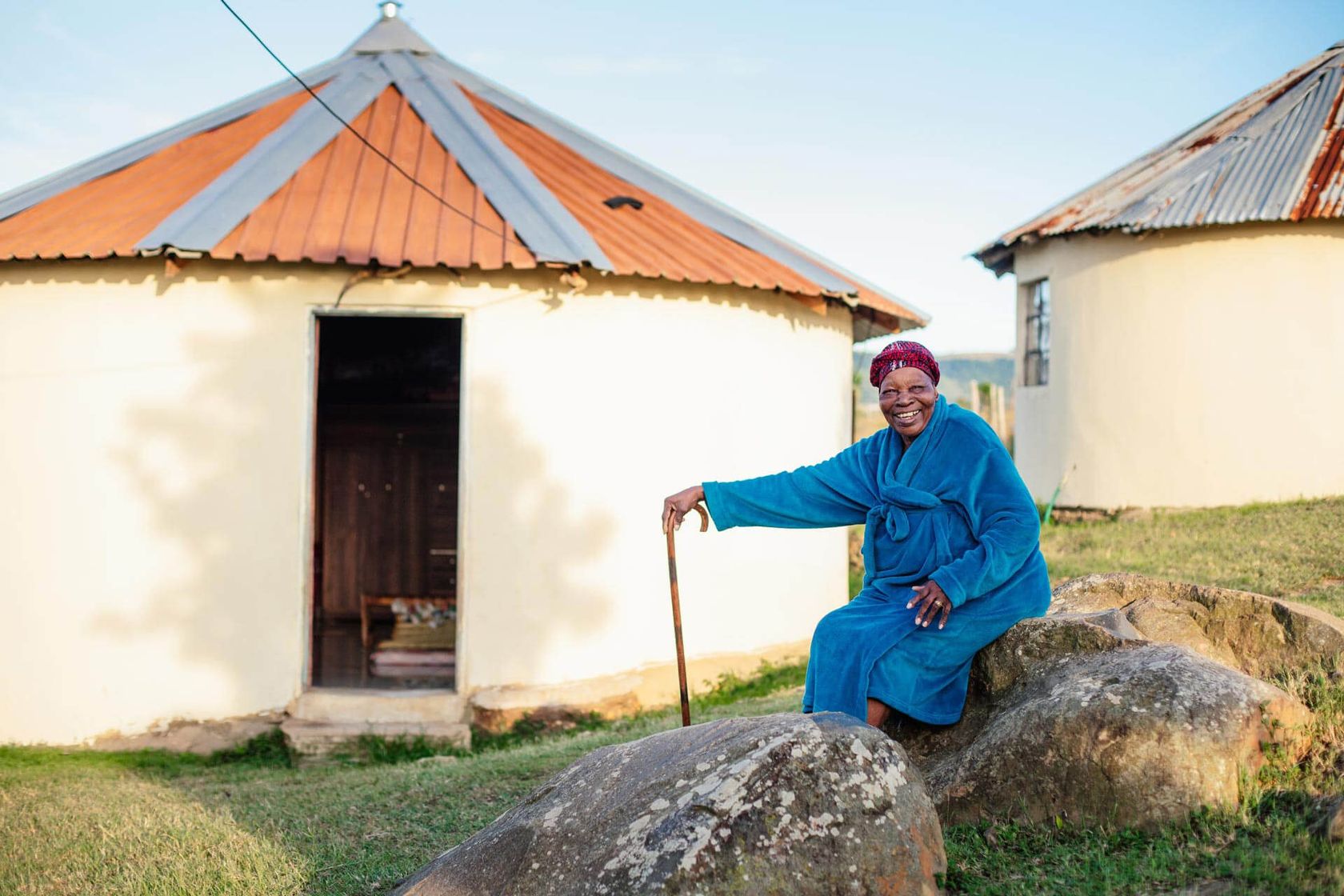 A community elder in a blue traditional dress sits with a cane on a rock before a thatched rondavel.