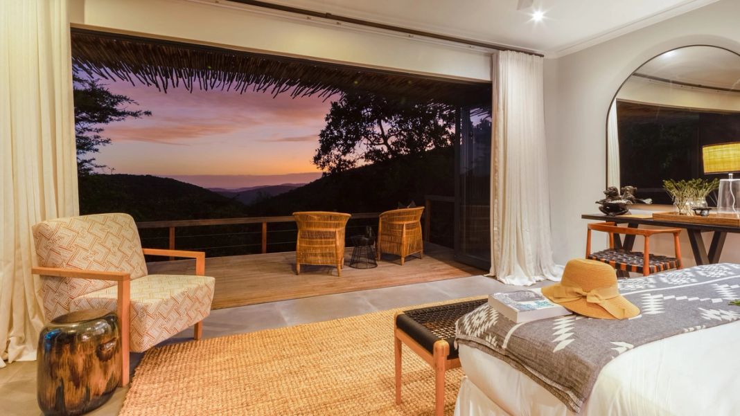 An open suite looks onto a deck with two chairs and a pink dusk sky over layered hills beyond.