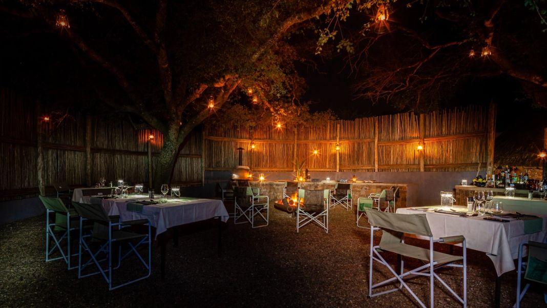 Tables with white linen and director chairs stand on gravel under a lantern-hung tree in a reed-walled boma at night.