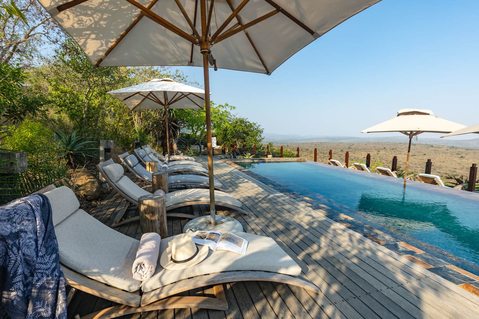 Cushioned loungers sit under white umbrellas beside the infinity pool, with the bushveld beyond.