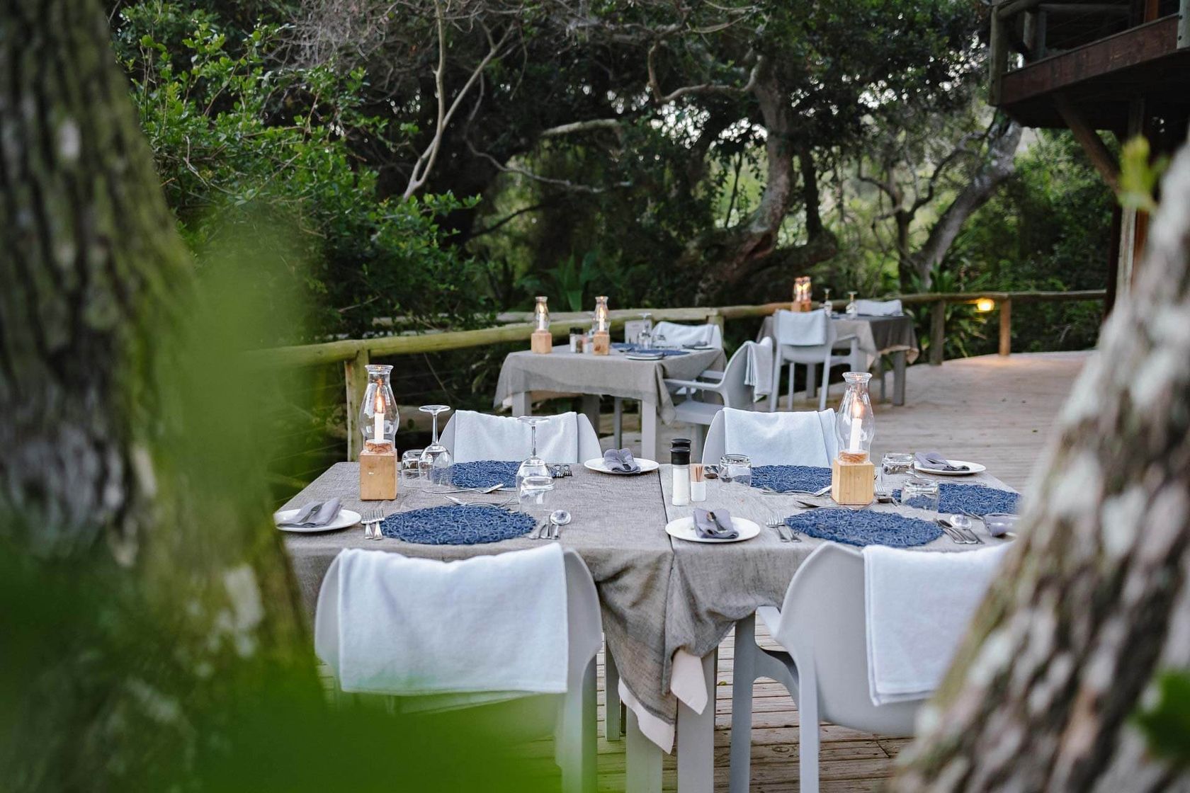 Outdoor deck tables set with grey linen, blue placemats and hurricane lanterns, viewed through trees beside a forest.