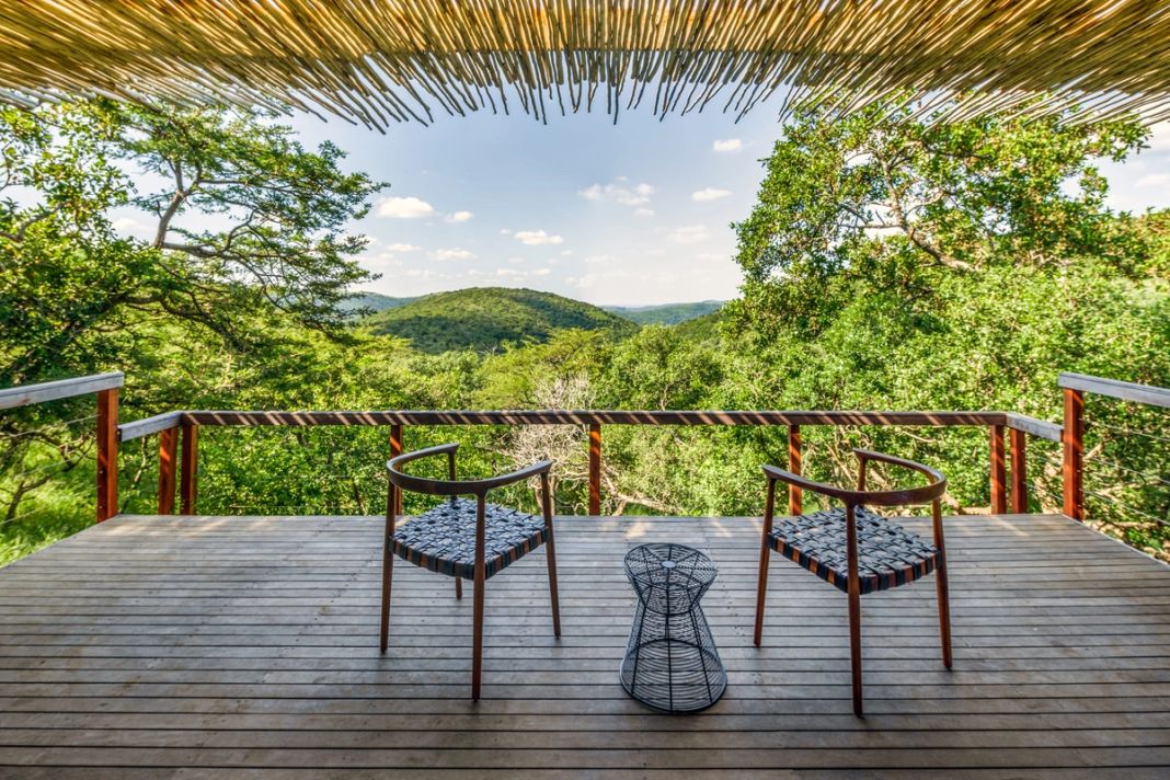 A suite deck under thatch with two woven timber chairs and a wire table framing a view of green hills.