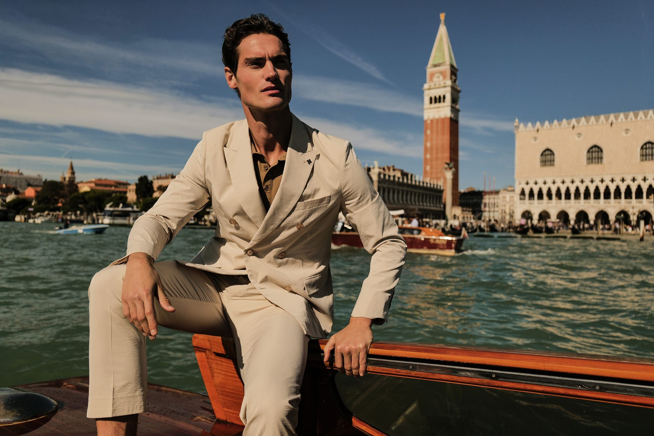 Distretto12 campaign: model in a cream double-breasted suit on a boat on the Venice lagoon, St Mark's Campanile behind