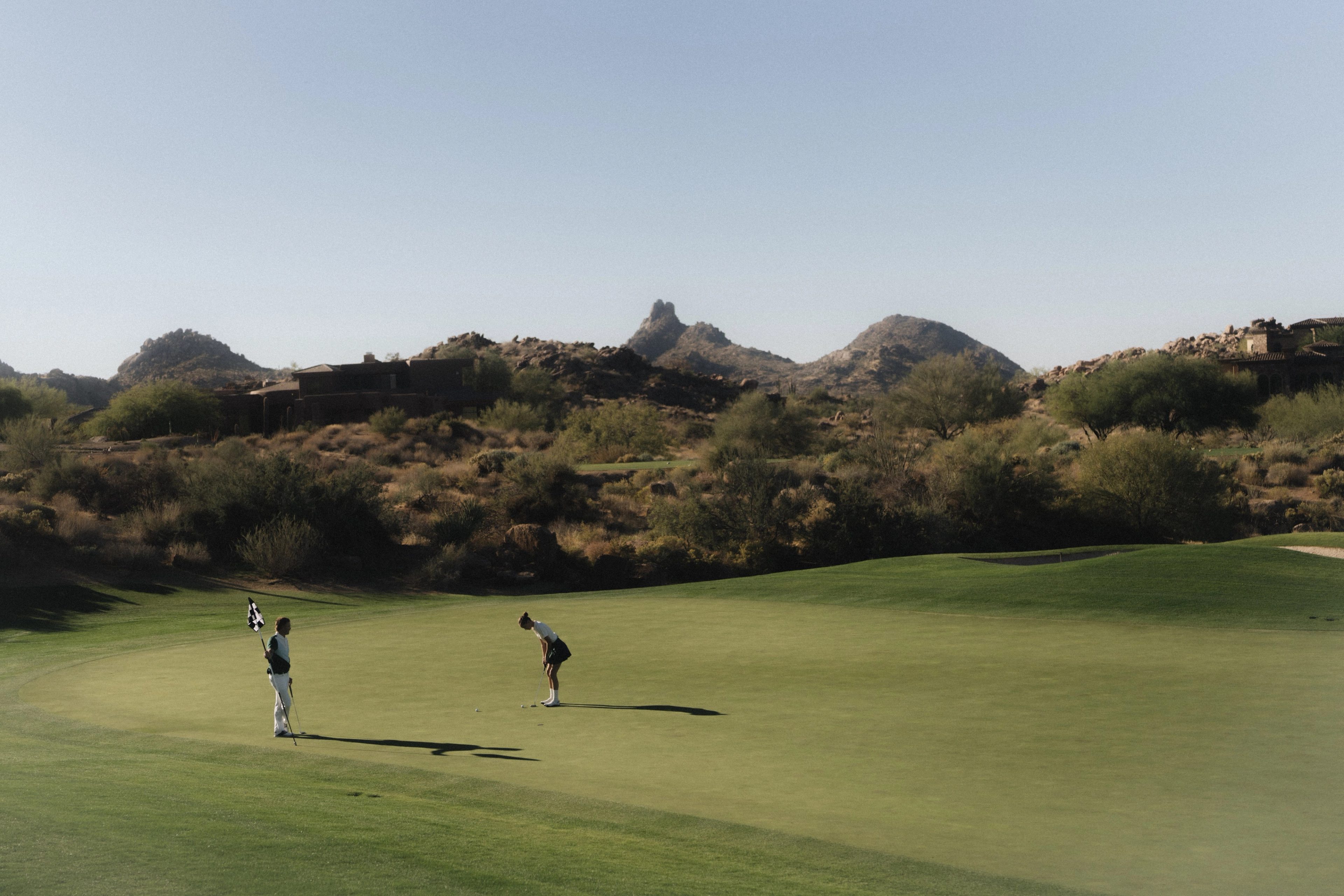 Two golfers on a putting green at a desert course, rock formations behind