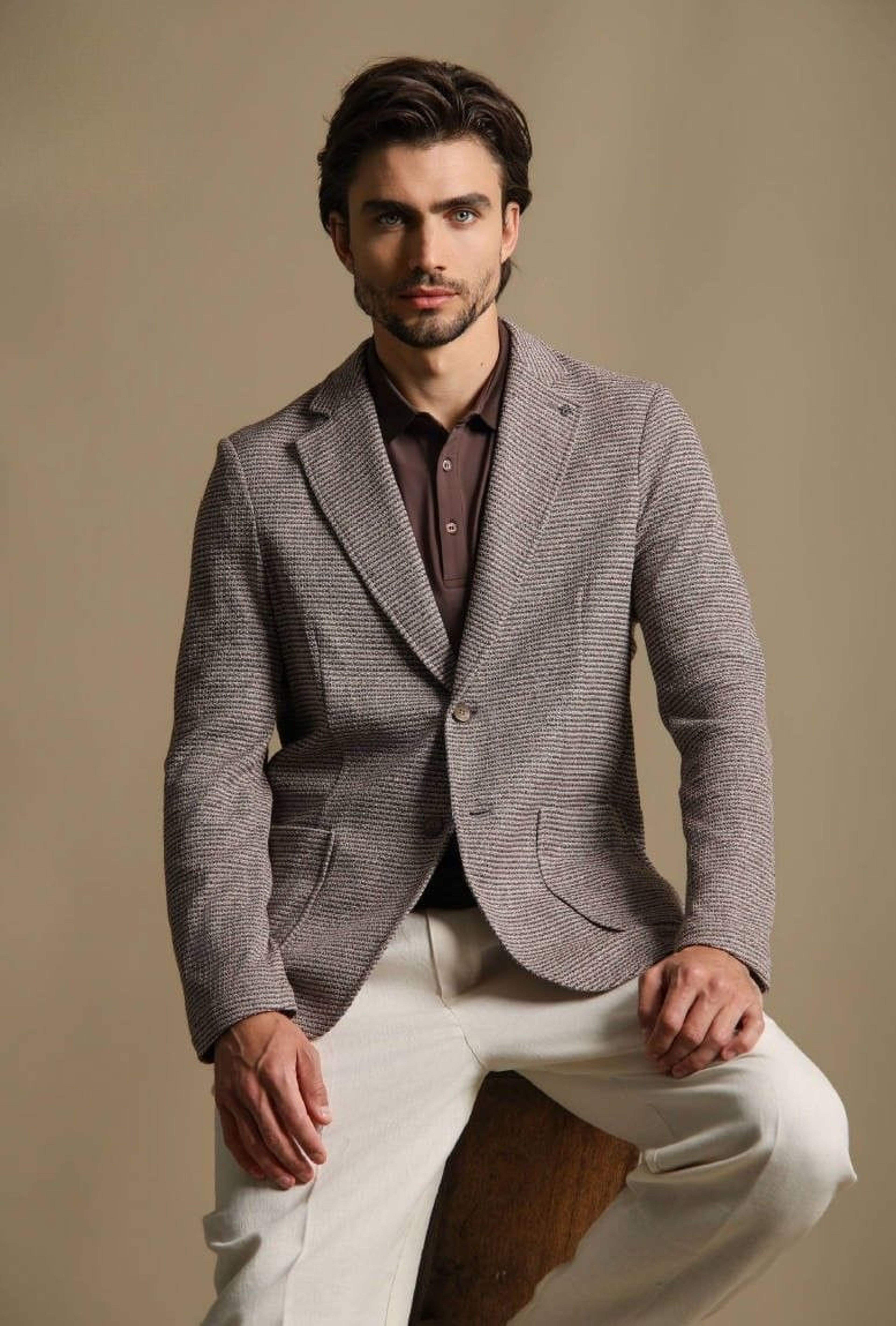 Model in a Distretto12 knitted tweed blazer over a brown knit polo, seated against a beige backdrop