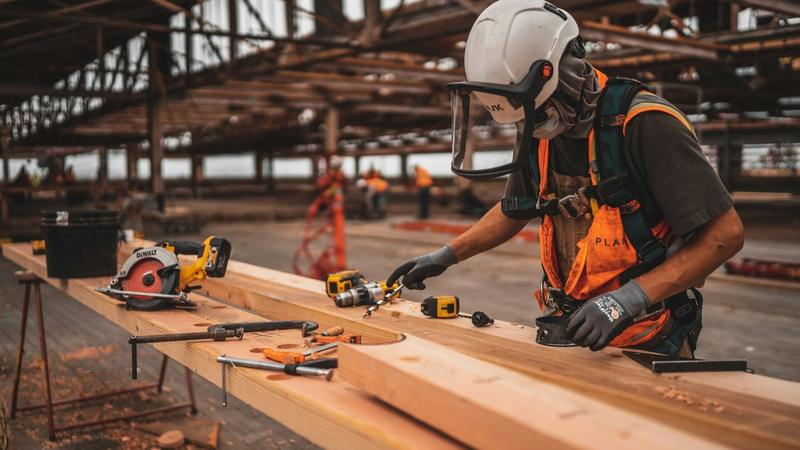 A maintenance technician in a hi-vis vest and hard hat operates a power tool on an indoor building job.
