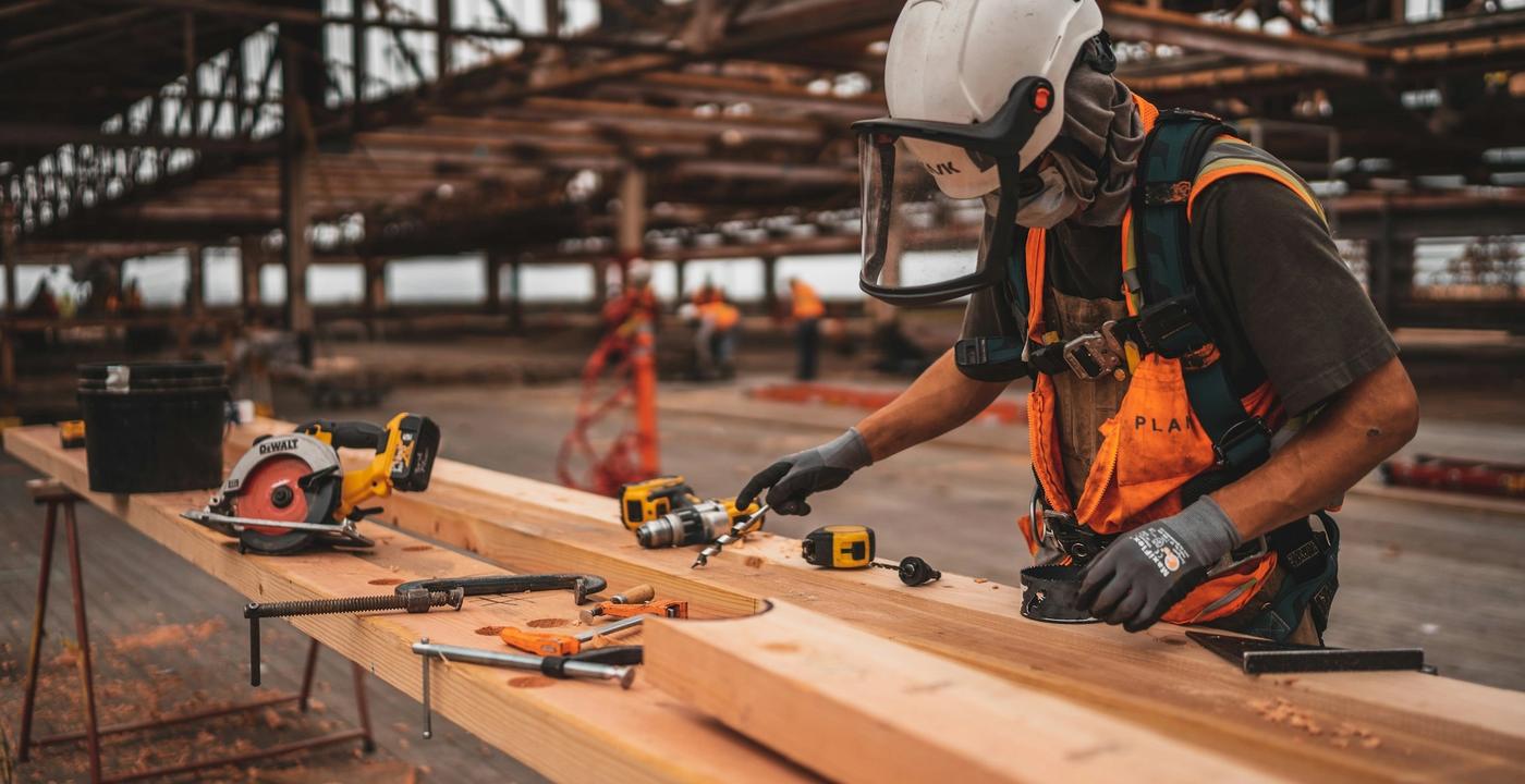A maintenance technician in a hi-vis vest and hard hat operates a power tool on an indoor building job.