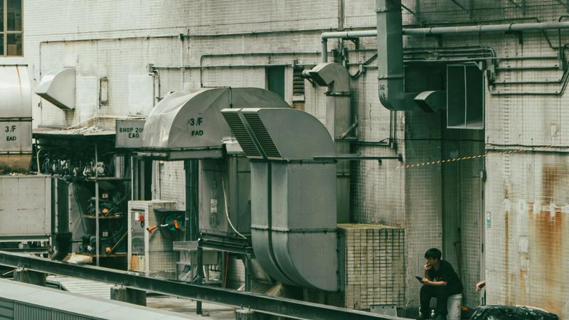Two workers rest in a rooftop mechanical area surrounded by HVAC units and ducting on an urban building.