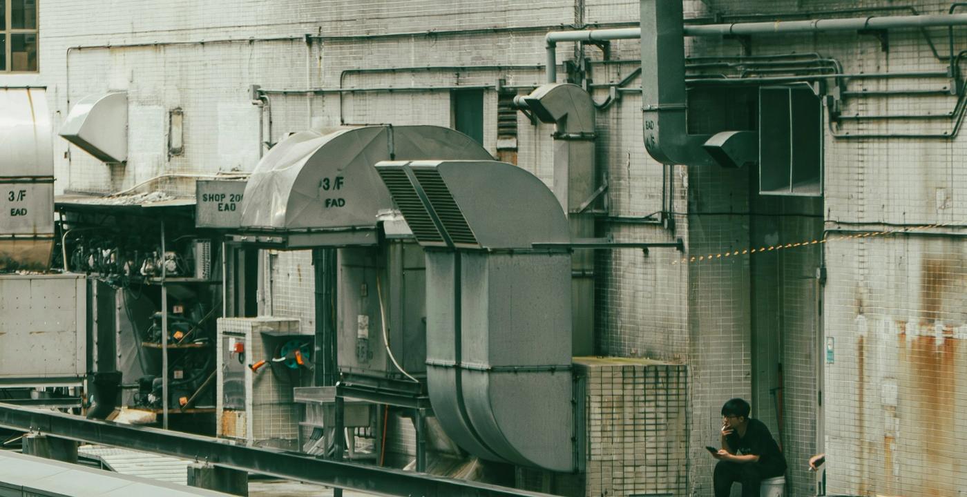 Two workers rest in a rooftop mechanical area surrounded by HVAC units and ducting on an urban building.