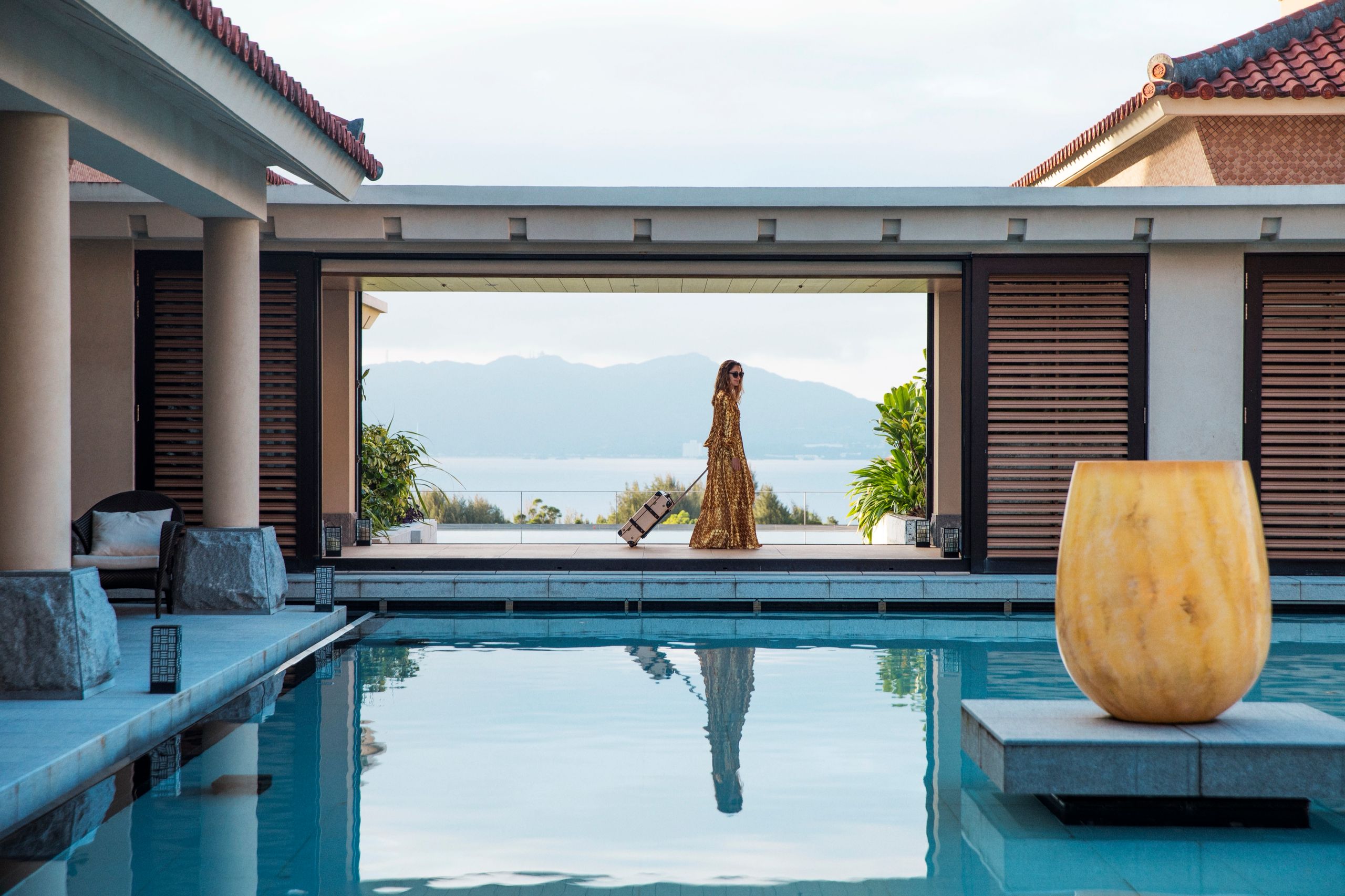 A woman in a gold gown wheels a suitcase past an infinity pool at a villa overlooking the sea in Okinawa.