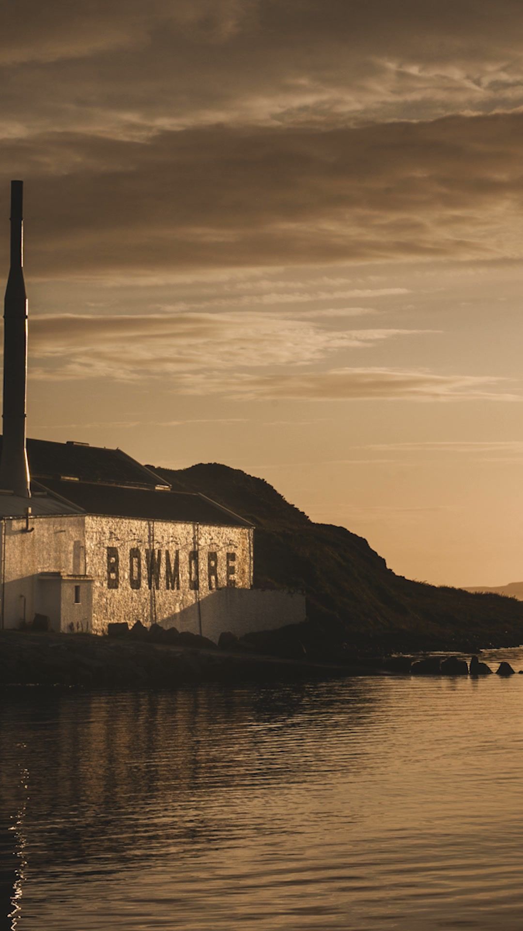 The Bowmore distillery on the Islay shoreline at golden hour, its name painted across the whitewashed warehouse wall above the sea loch.