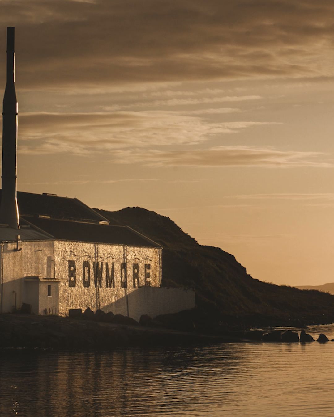 The Bowmore distillery on the Islay shoreline at golden hour, its name painted across the whitewashed warehouse wall above the sea loch.