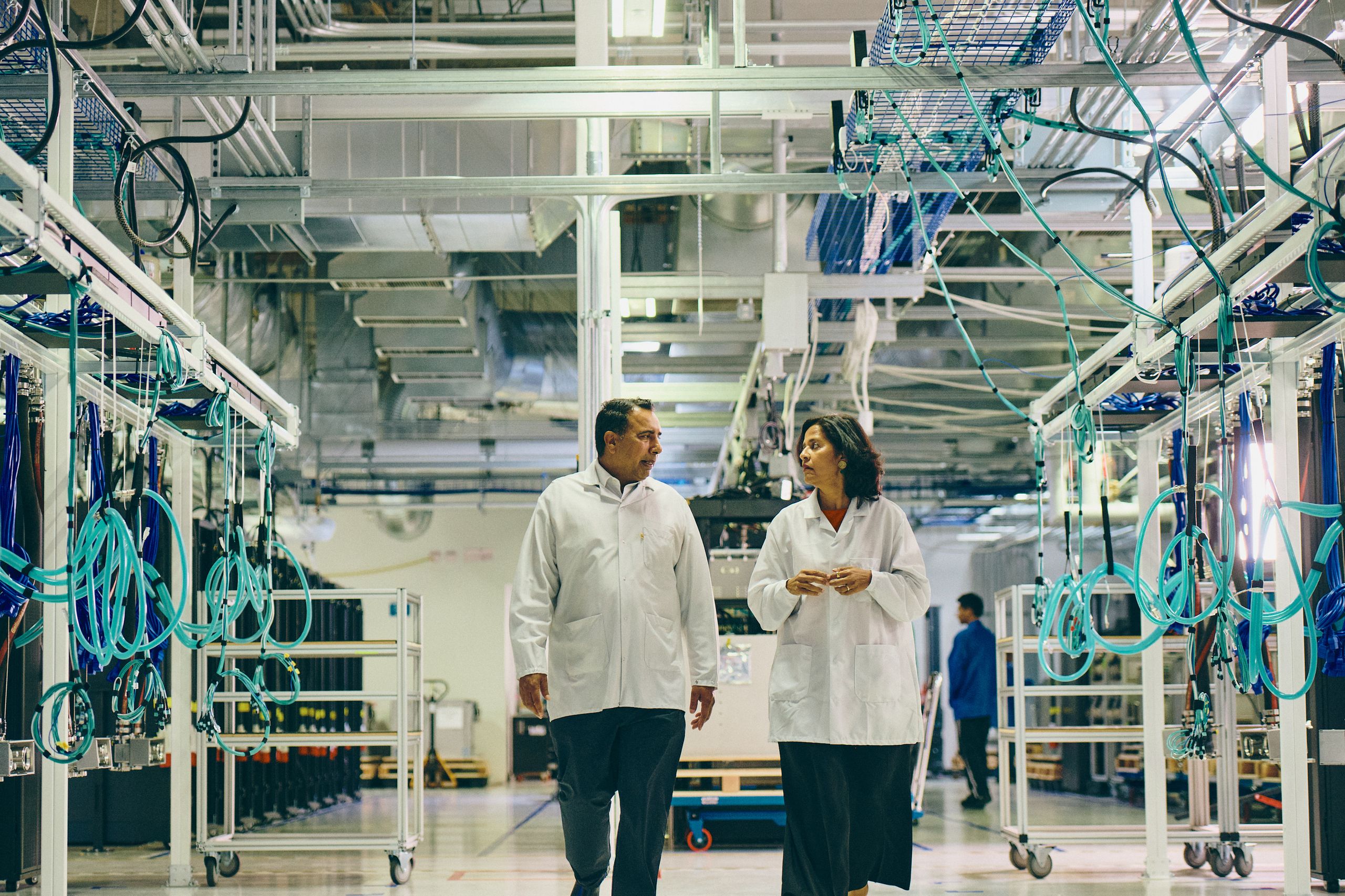 Two engineers in white coats walking and talking down an aisle of the Axiom production floor, between racks strung with teal cabling