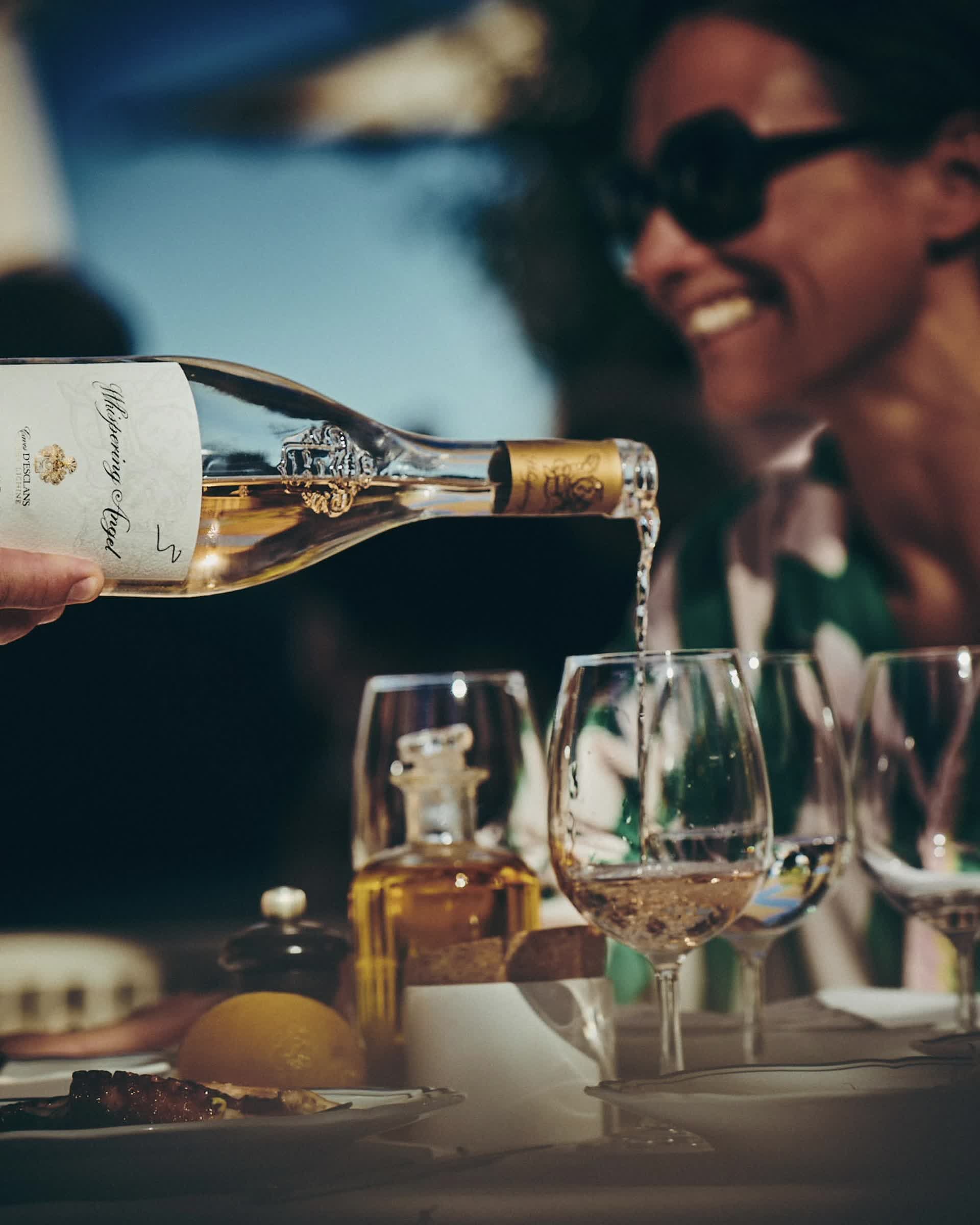 A bottle of Whispering Angel rose being poured into glasses at a sunlit table, a guest laughing in the background