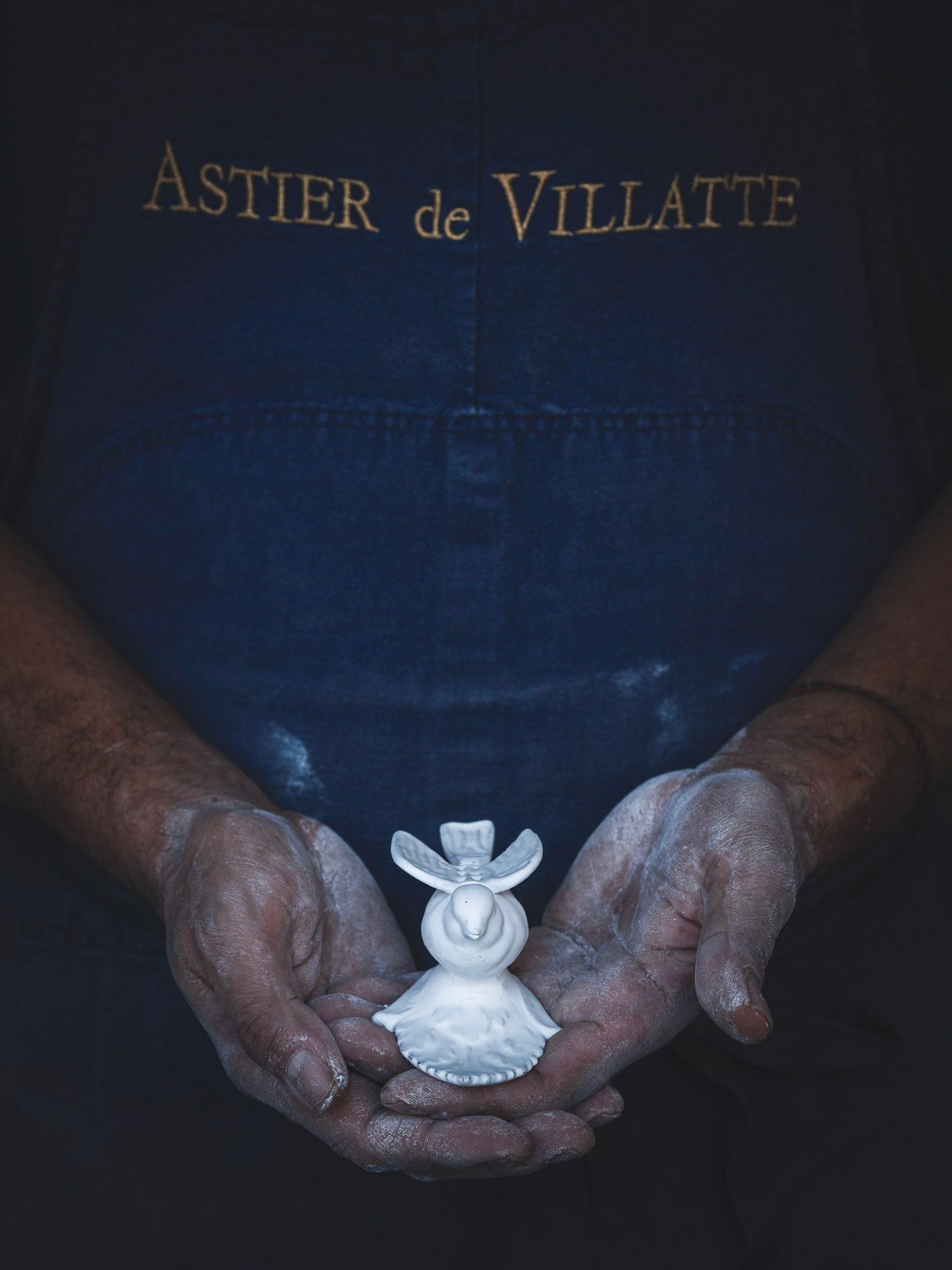 A potter's clay-dusted hands cradling a small white Astier de Villatte ceramic bird figurine