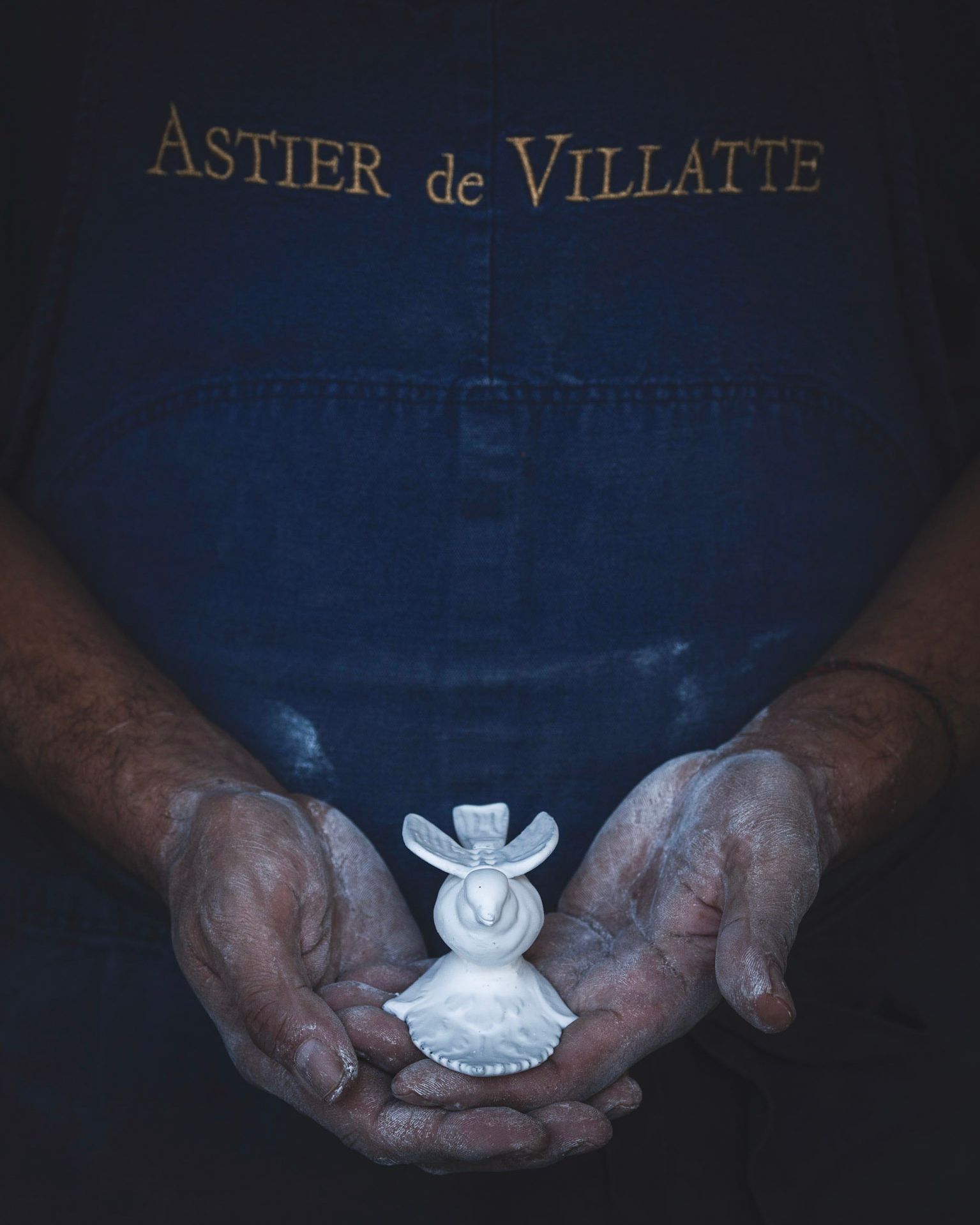 A potter's clay-dusted hands cradling a small white Astier de Villatte ceramic bird figurine