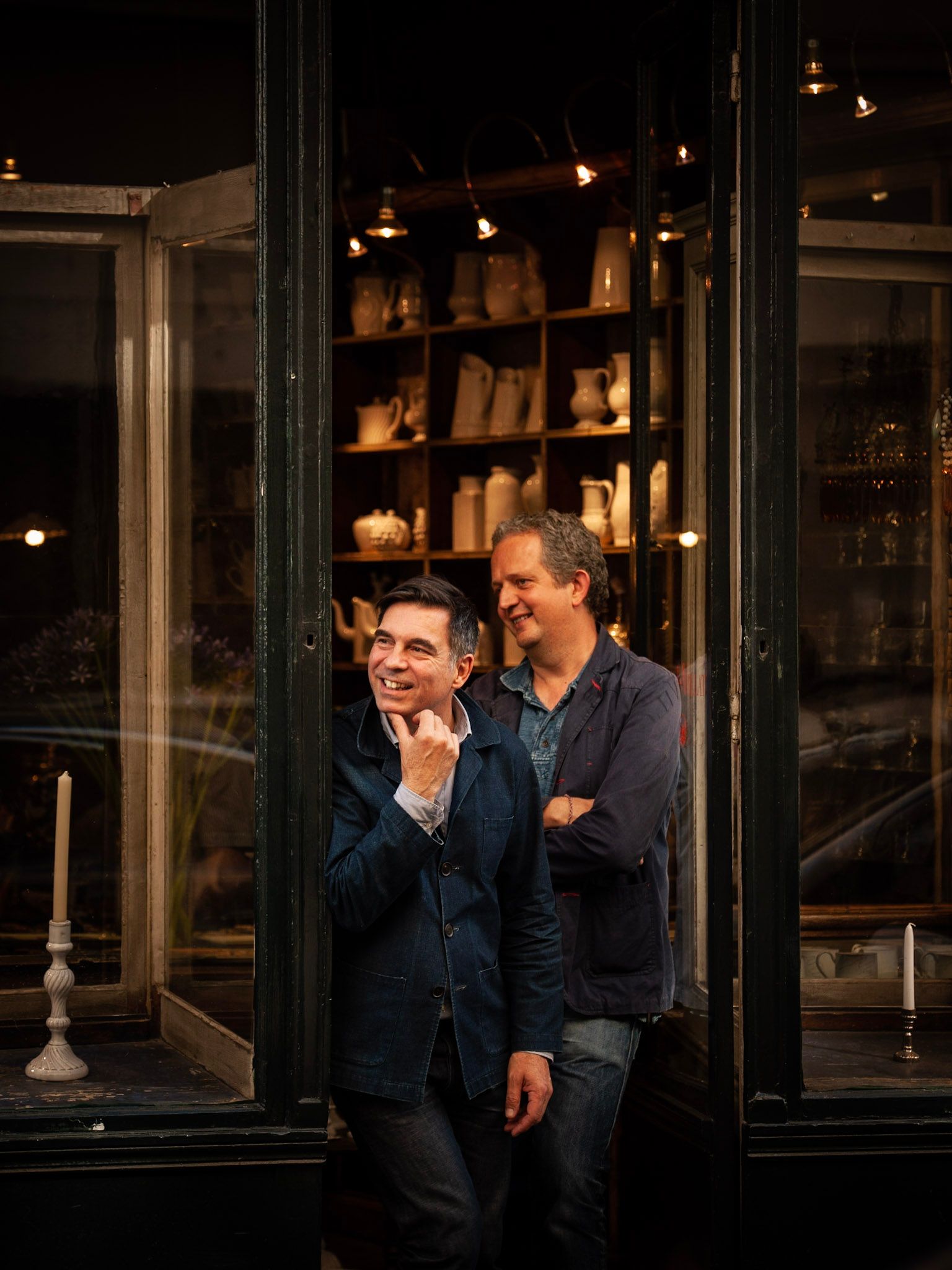 The founders of Astier de Villatte in the doorway of their Paris shop, shelves of white ceramics behind them