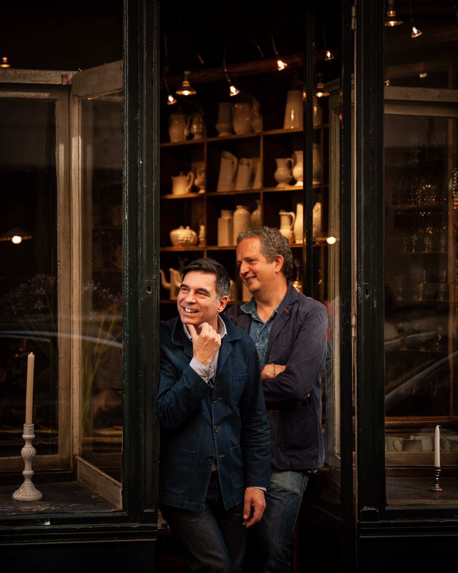 The founders of Astier de Villatte in the doorway of their Paris shop, shelves of white ceramics behind them