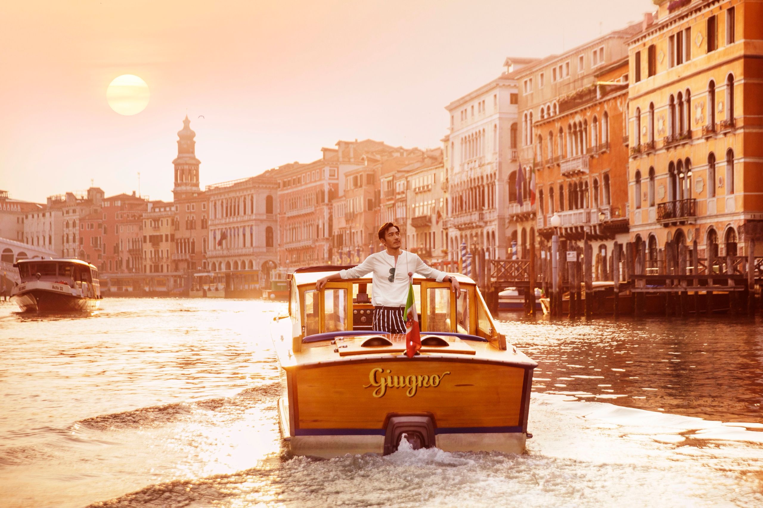 A man rides the bow of a wooden water taxi along the Grand Canal in Venice at sunset.