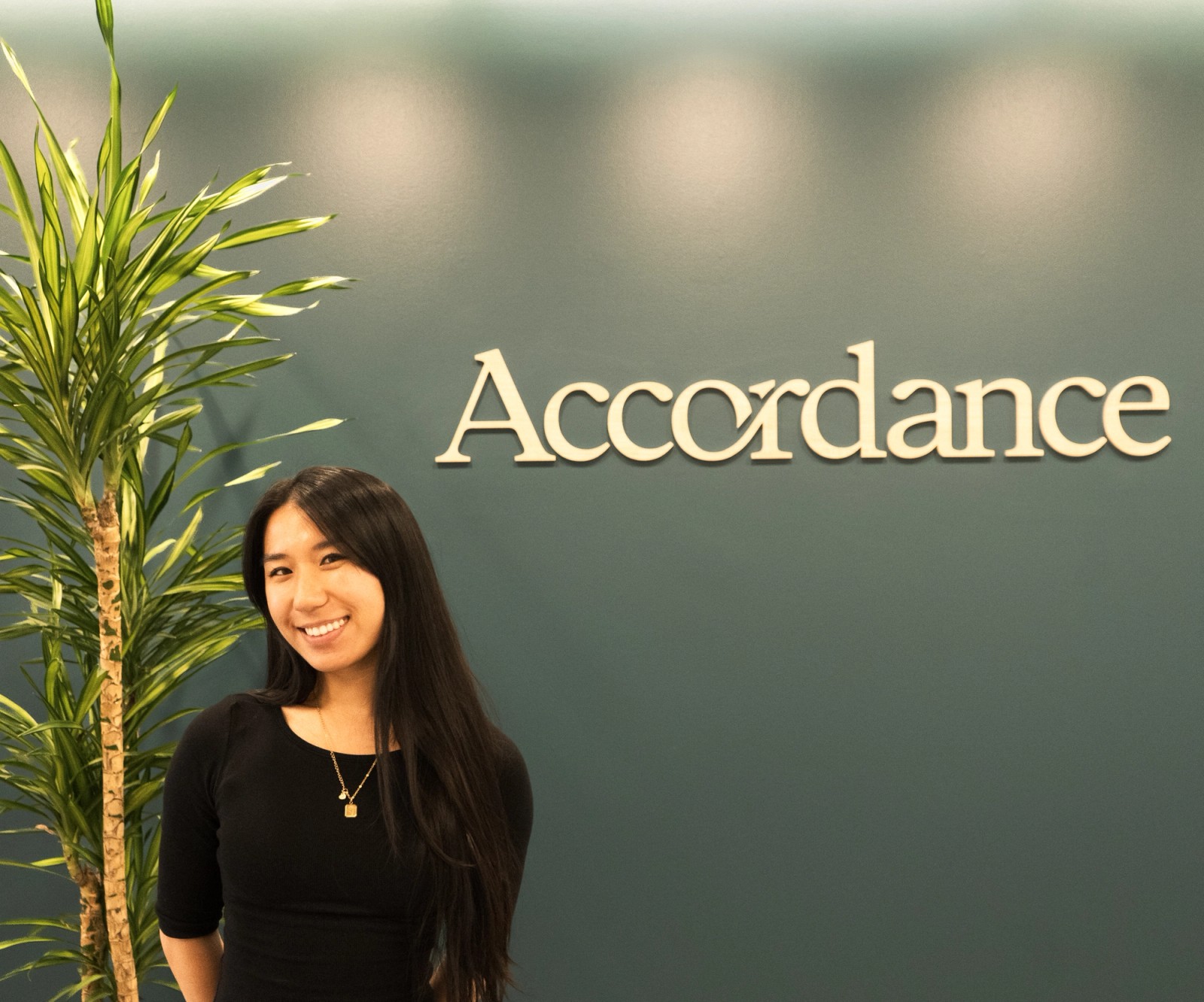 Smiling woman standing by a plant, with the word "Accordance" on the wall behind her.