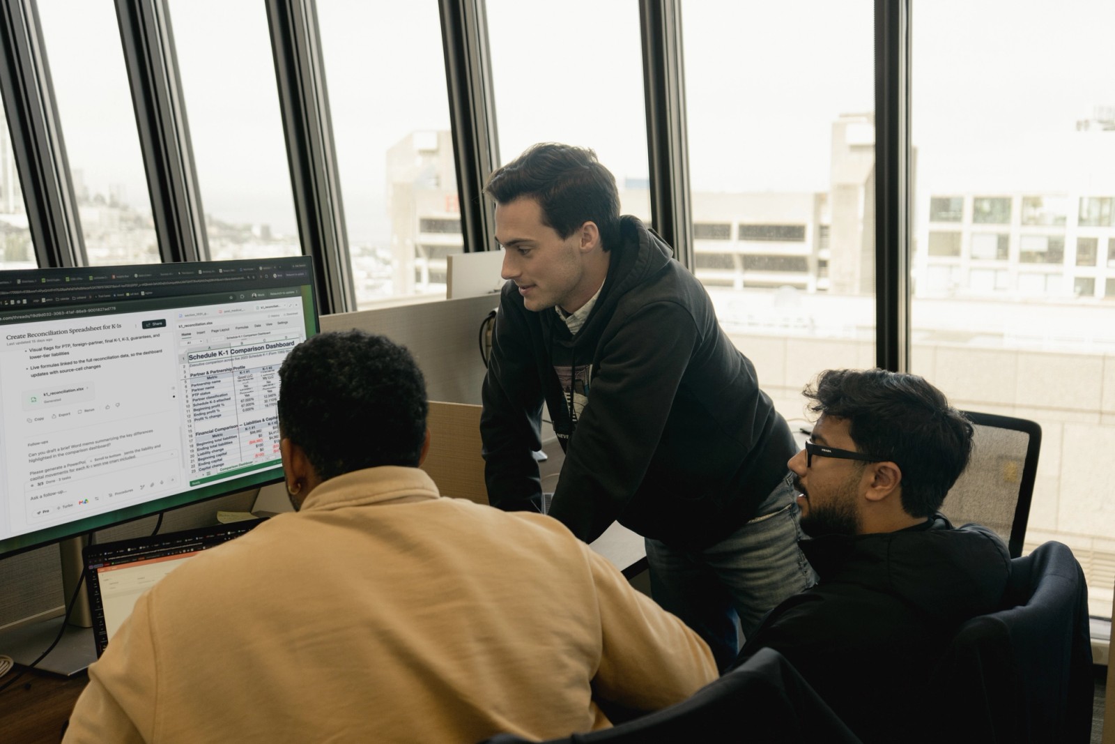 Three men collaborate around a computer monitor in an office with a city view.