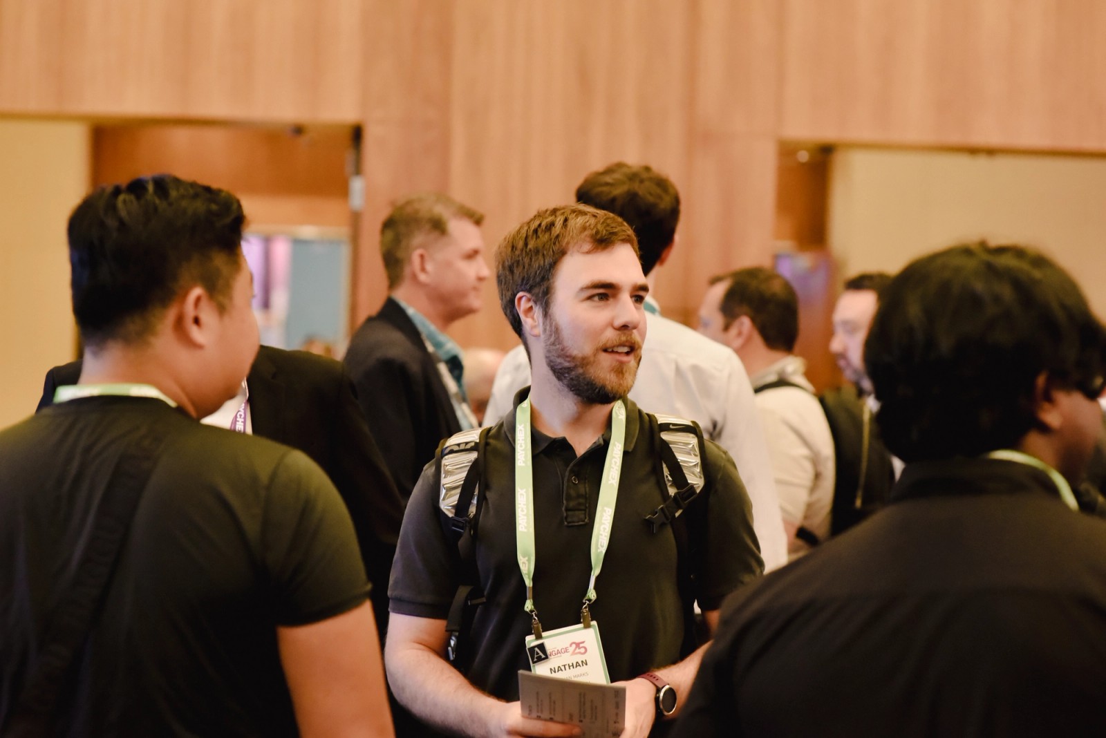 A man with a beard and an event badge talks with other attendees at a busy indoor event.