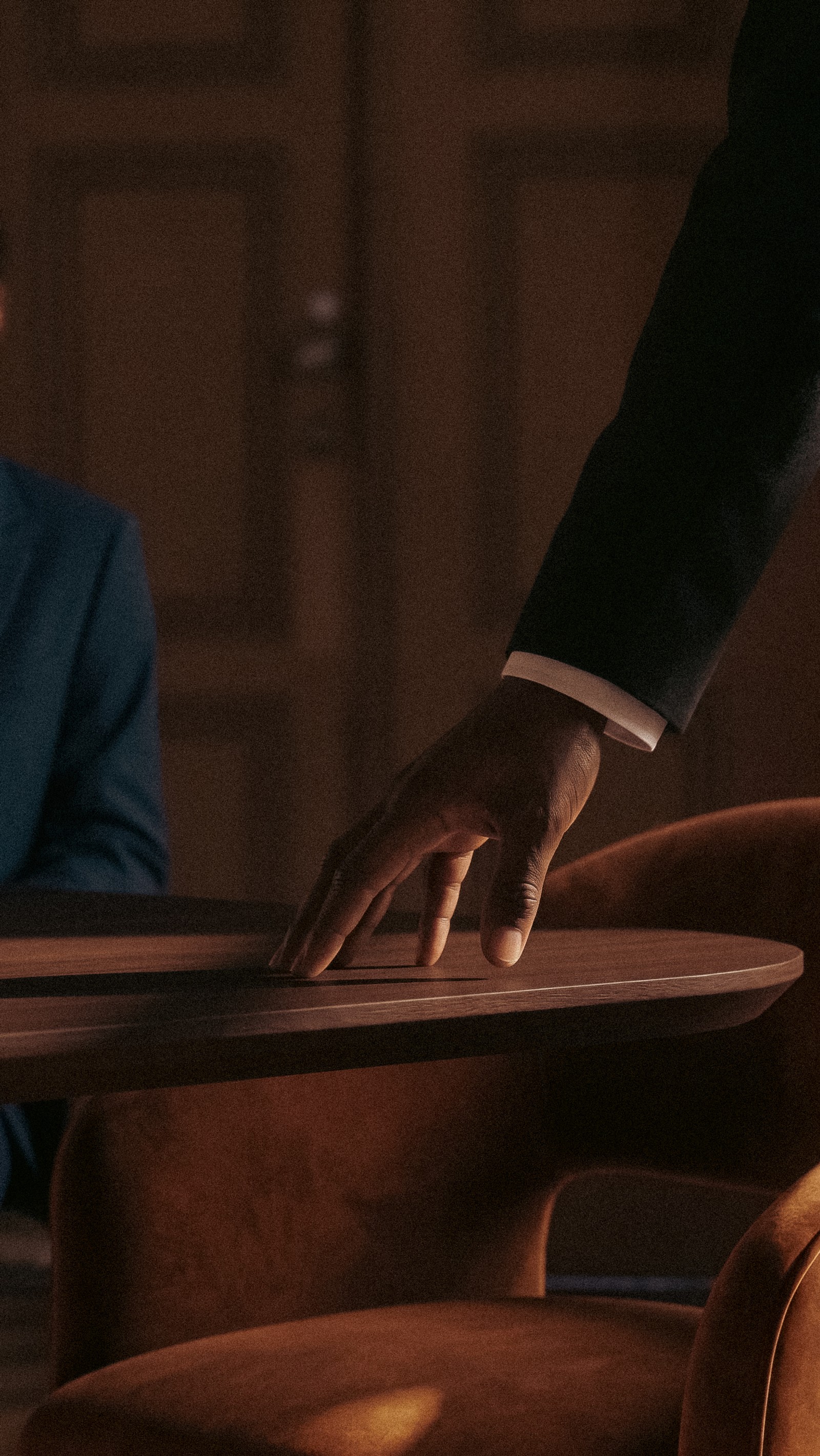 A dark-skinned hand in a suit rests on a wooden table, with a blurred person in a blue jacket in the background.