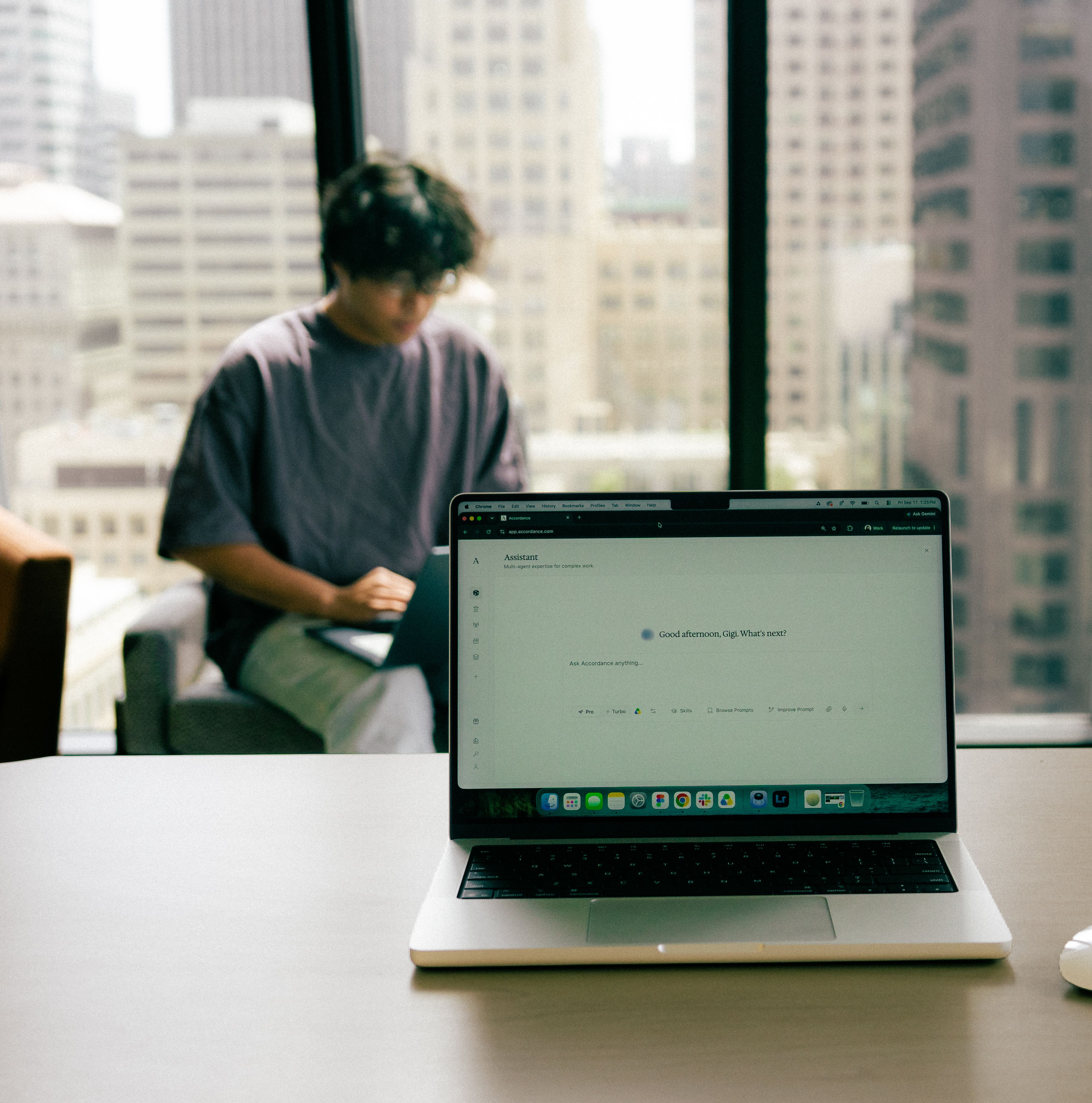 A laptop displaying an AI prompt, with a blurry person using another laptop and a cityscape in the background.