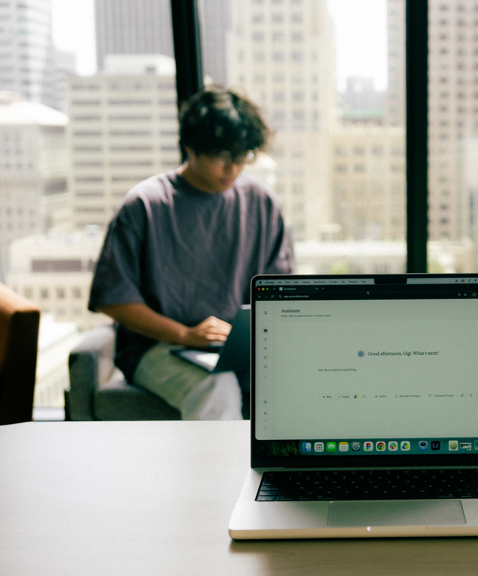 A laptop displaying an AI prompt, with a blurry person using another laptop and a cityscape in the background.