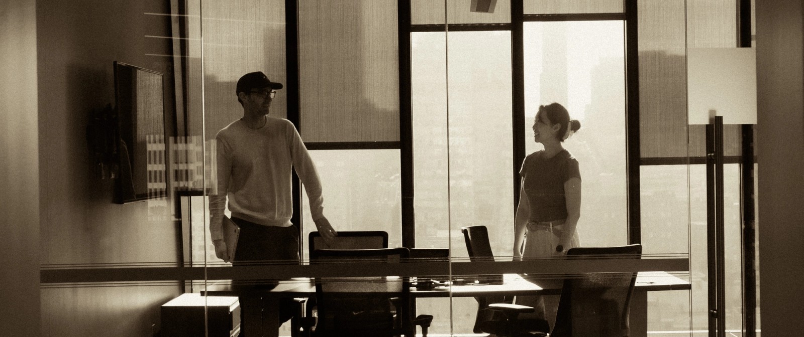 Sepia-toned image of two people standing in a bright conference room with large city windows.